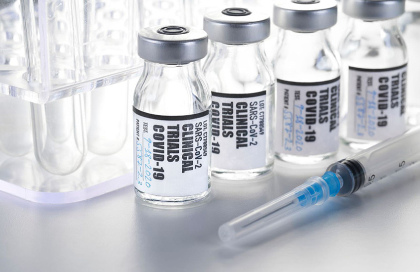 vials labeled "clinical trials COVID 19" in front of test tube rack, with syringe in foreground