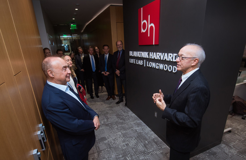 Two men in suits stand in front of a sign with a red square logo with letter B and H above white type on black background that reads Blavatnik Harvard Life Lab Longwood