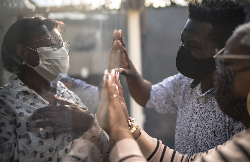 A woman on one side of a window faces a man and a woman on the other side, all three wearing protective masks.