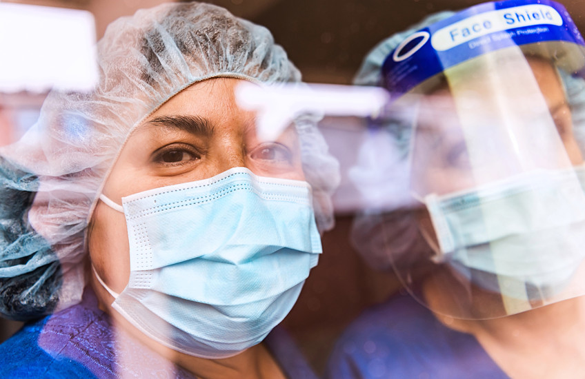 The faces of two people in medical scrubs, protective masks, and head gear seen through a reflection-covered window.