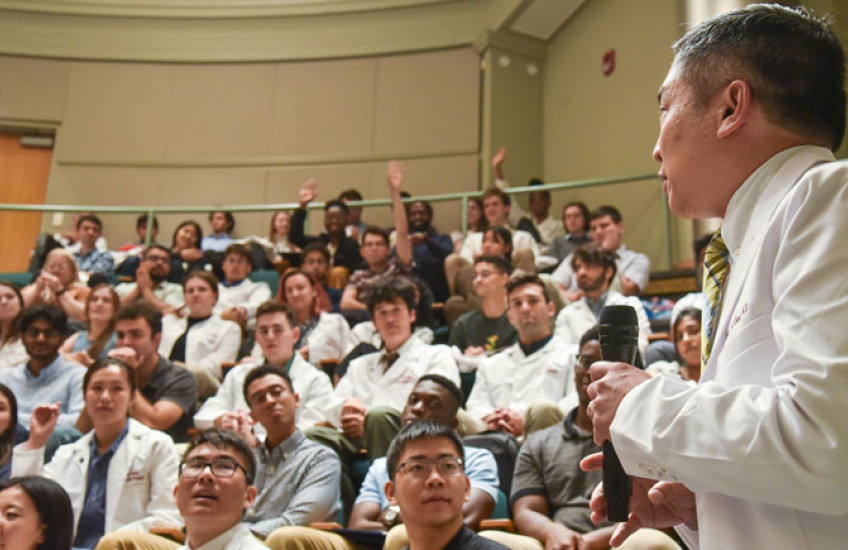Sideview of Chang in white coat holding mic while speaking to audience of students, many in white coats