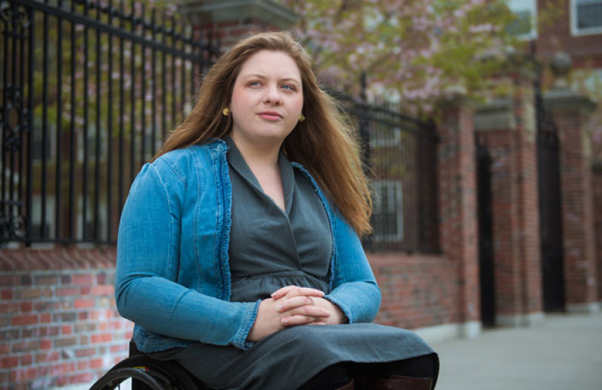 Cloutier sitting in wheel chair with arms folded in lap wearing a green button down and aqua sweater