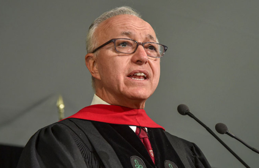 A gray-haired man with glasses and doctoral robes speaks into a microphone