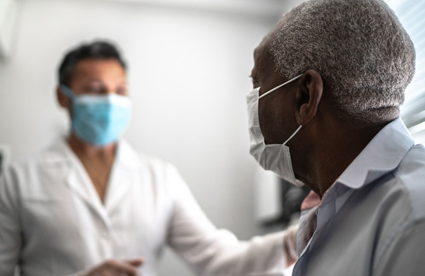 A dark-haired man in a white lab coat sits in a brightly lit room with a gray-haired man in a dress shirt.