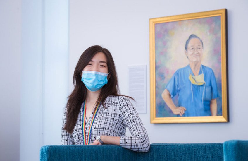 Photo of Pamela Chen, a young Asian woman wearing a mask and patterned shirt standing proudly in front of the portrait she painted of Yeu-Tsu Margaret Lee, who is an older Asian woman in the portrait looking determined while wearing her hair in a bun, scrubs, and a medical mask around her neck