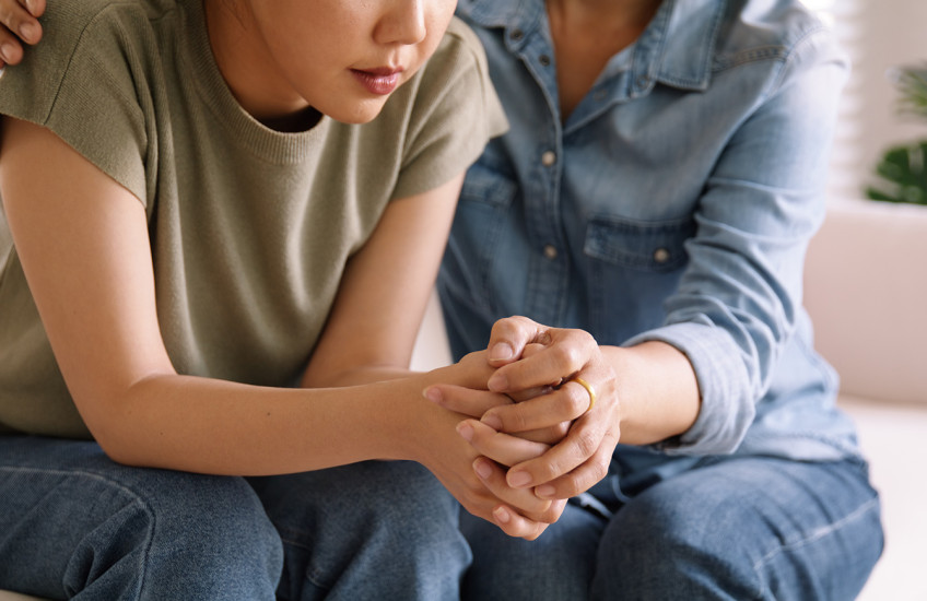 An older person holds a distressed younger person’s hands and shoulder, offering comfort.