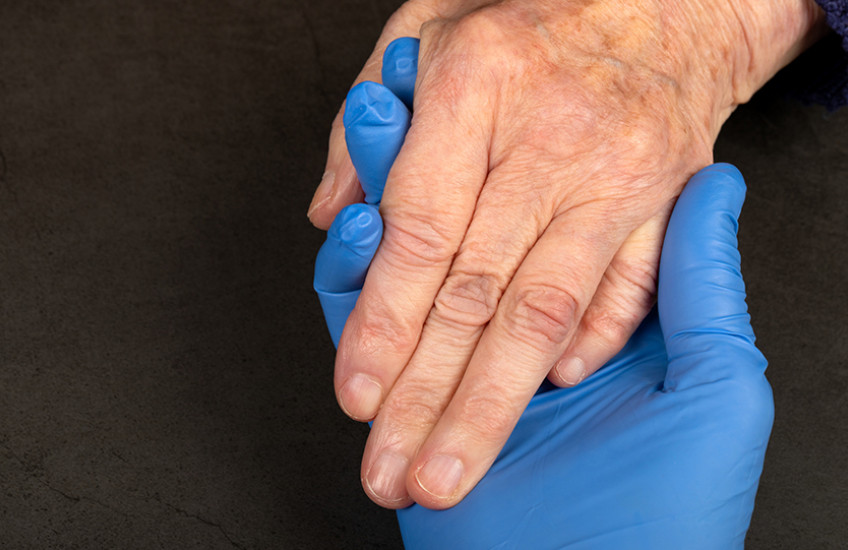 A hand in a blue latex glove holds the ungloved hand of an older white person