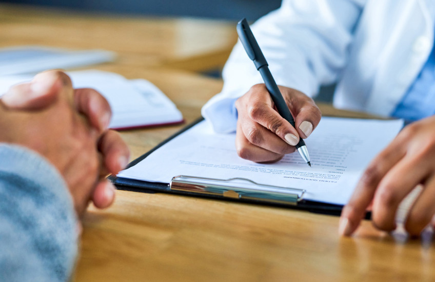 Photograph of doctor's hand writing on a form across from patient with hands folded
