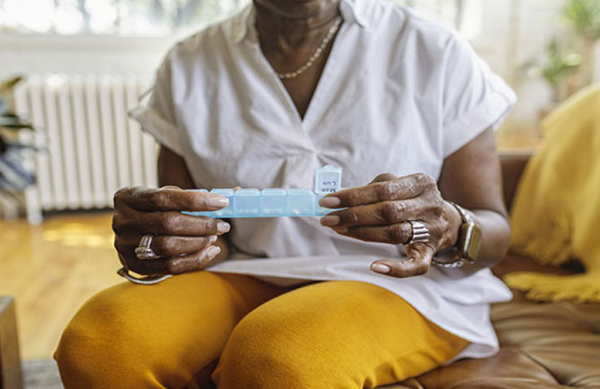 Woman sitting on a chair holding a pill case; wearing white blouse and gold pants