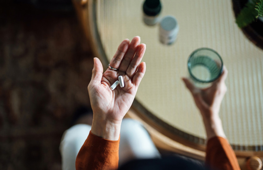 Photo looking down to a woman's hands, one holding a glass of water, in the other hand, some pills