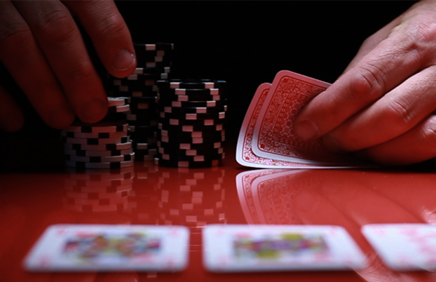 an unseen person at a shiny gambling table holds unknown cards while picking up a few poker chips from a stack. on the table lie three blurry cards face-up.