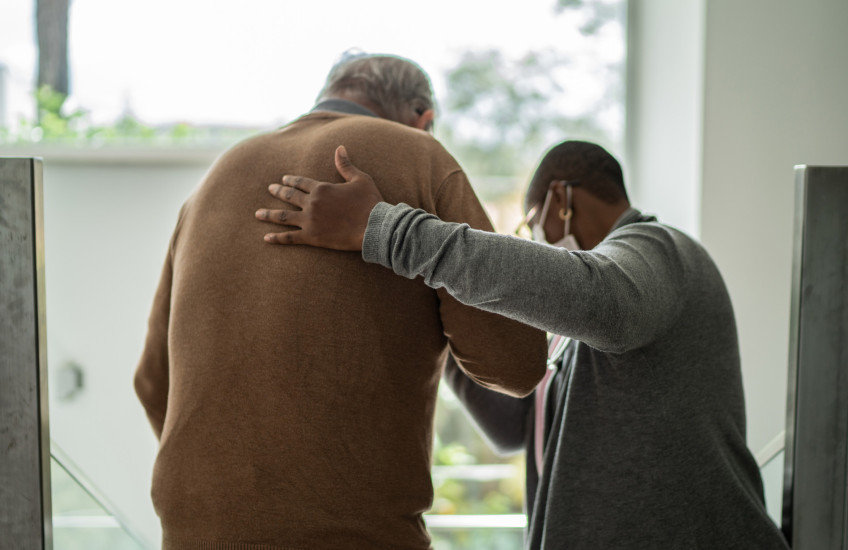 Rear view photo of health care worker helping a patient to walk, inside a room with windows