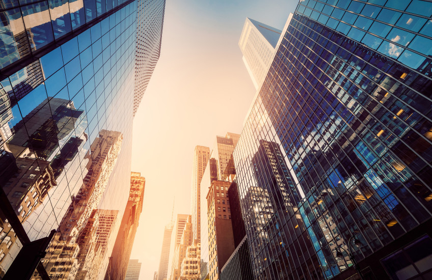 Photograph of tall office buildings, taken from the street, looking up to a sunny sky and tops of skyscrapers