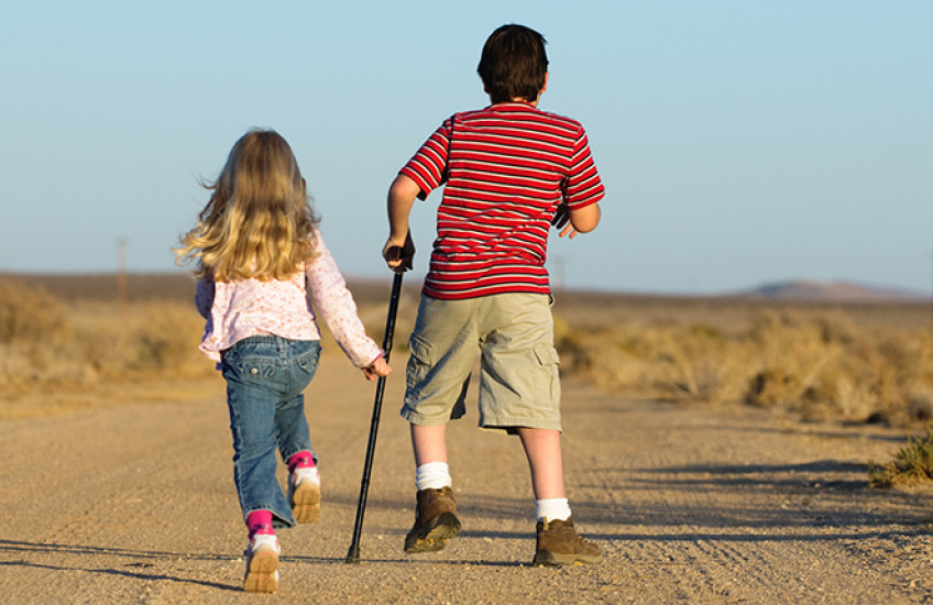 Two children - one girl unassisted and one boy with a crutch - skip down a dirt road