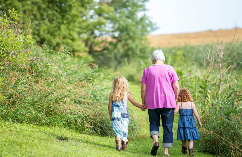 Woman in pink top walking, holding hands with two little girls, backs to camera, in a garden