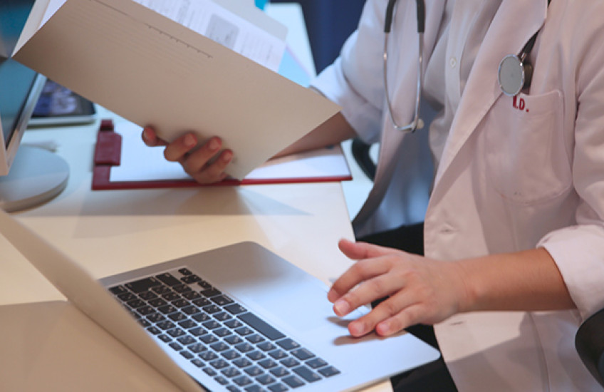 Photo of physician holding a pen and a notebook and typing on a computer keyboard