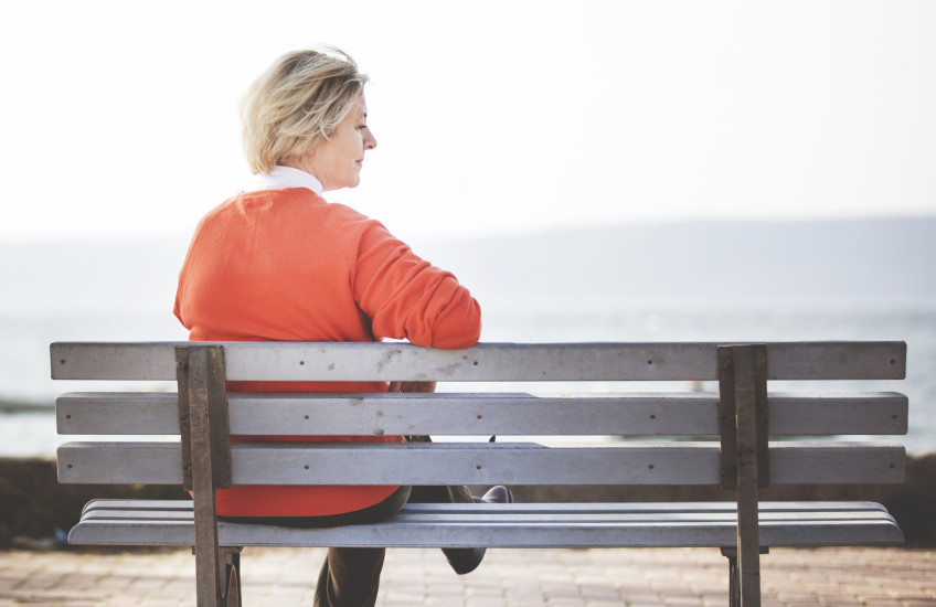 Woman with light hair and orange sweater sits alone on a bench overlooking water