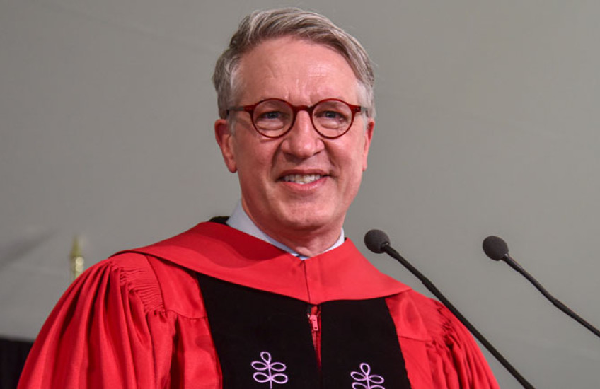 A white man in glasses and red robes smiles at a microphone