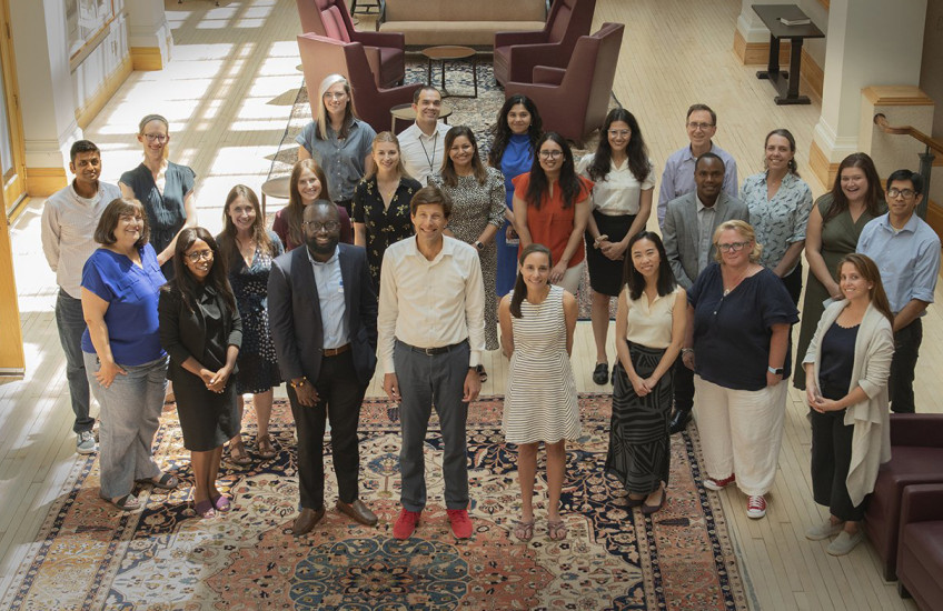 Photographed from above, 24 people stand in the center of a sunlit room smiling up at the camera.