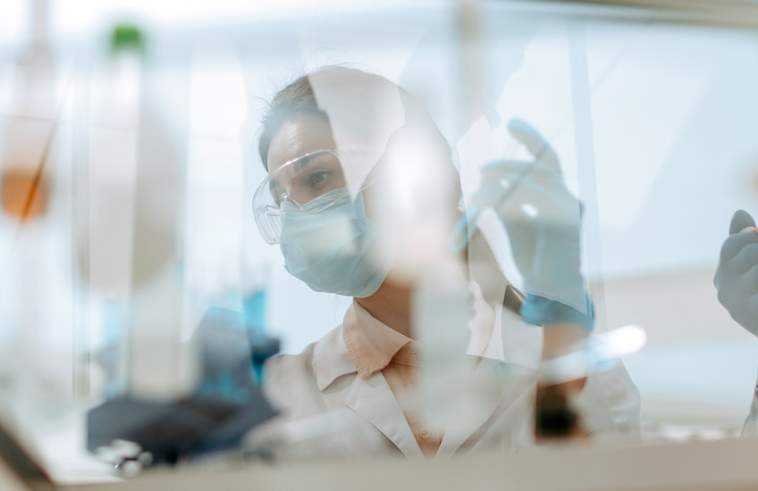 A woman in safety glasses, mask and lab coat working at a laboratory table seen through the reflections and refractions on a safety glass window.