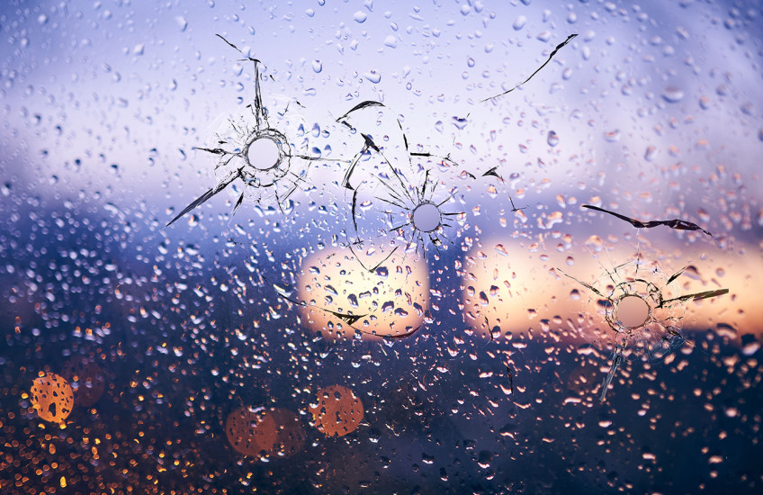 City lights reflected in a raindrop-speckled window, the glass shattered by several round bullet holes with jagged cracks.