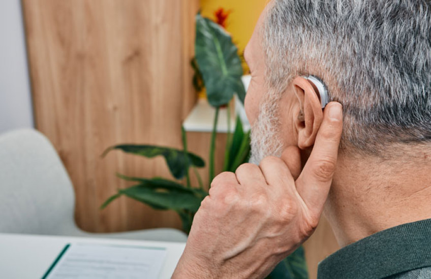 back of the head of a white grey-haired man holding a finger to his ear that has a hearing aid