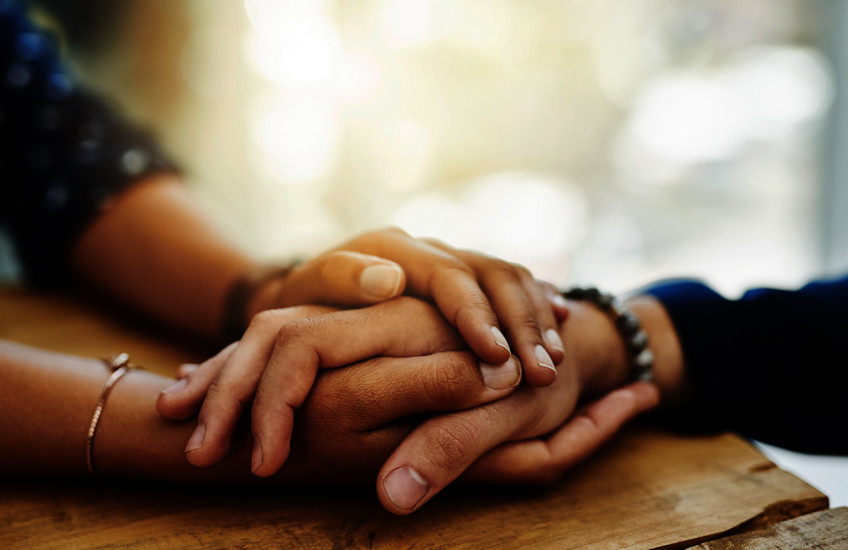 Closeup of two people’s hands clasped together on a table in a comforting manner.