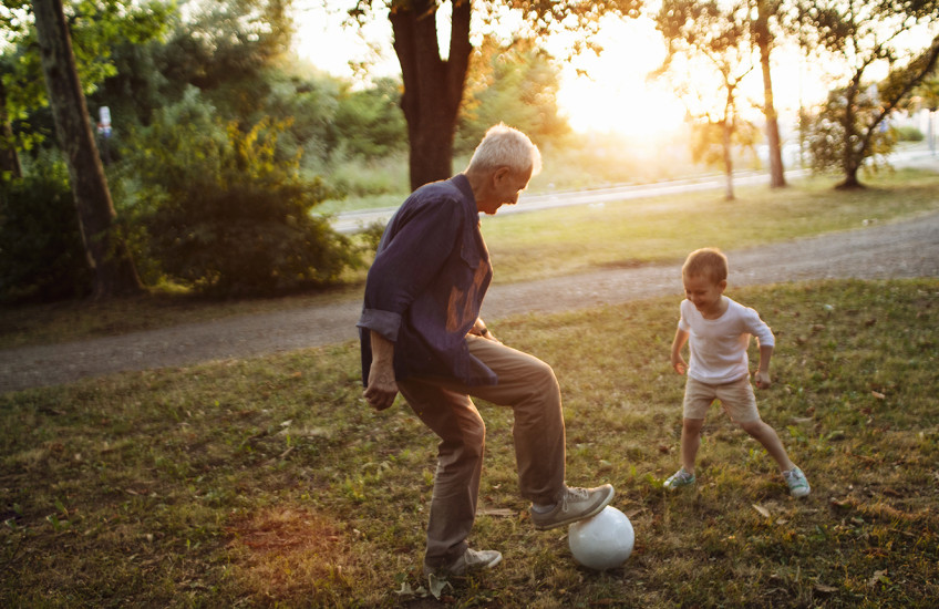 Caught in the middle of a one-on-one soccer game, a wite-haired man stands with his foot on a soccer ball, a small child faces him, poised to take the ball.