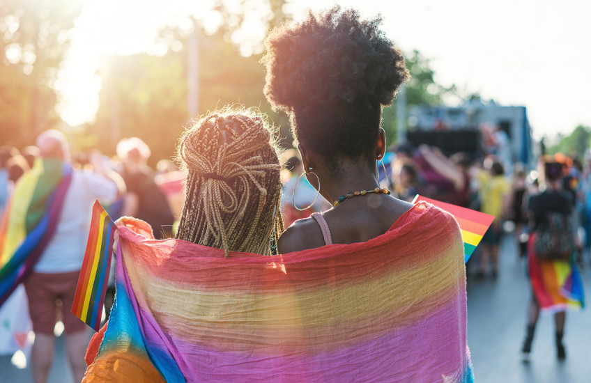 Rear view image of two people with braided, long hair, wrapped in a rainbow scarf walking with other people waving rainbow flags 