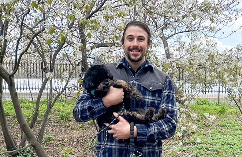 Nelson LaMarche with his miniature poodle in a NY city park with blooming pear tree and Hudson River behind him