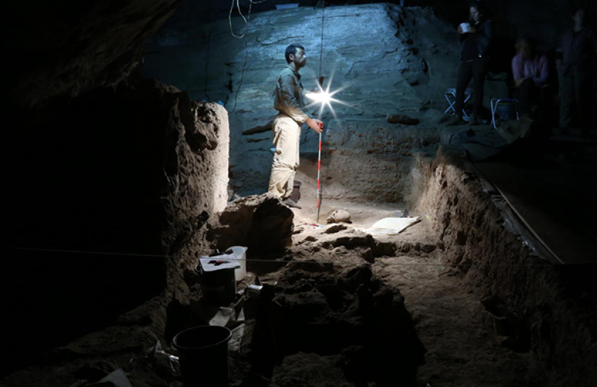 Man with lamp stands in a cave, an excavated skull in front of him