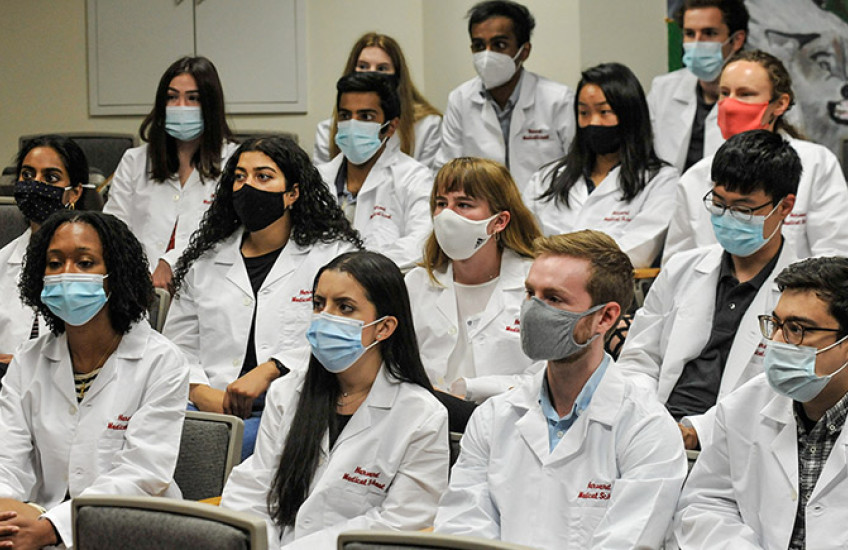 17 students wearing masks and in white coats that say Harvard Medical School seated in auditorium seats
