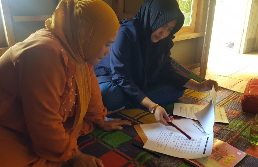 Two women, one in orange and one in blue seated on a colorful rug look at documents together.