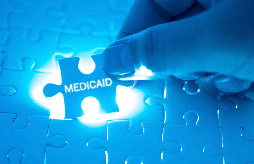 A hand fitting a puzzle piece adorned with the word "medicaid" into a back-lit jigsaw puzzle.