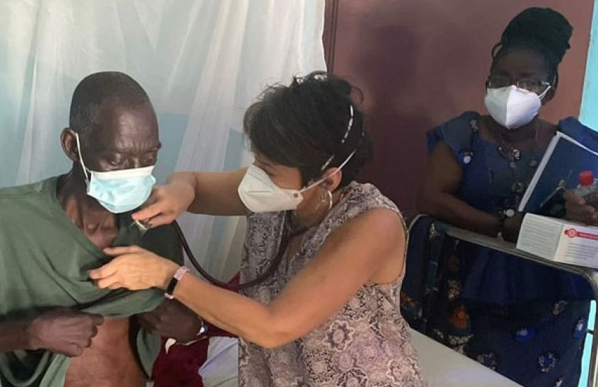 Mukherjee in a print dress wearing a mask holds a stethoscope to the chest of an older man who is wearing a mask while two masked women stand in the background