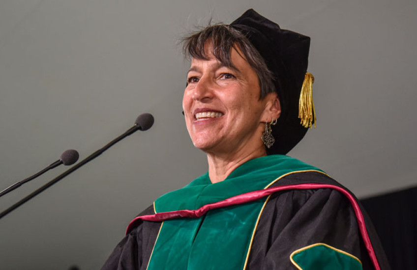 Close-up of a white woman with gray hair in doctoral robes smiling as she speaks into a microphone