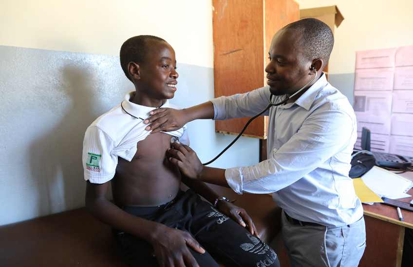 A teenage boy sits on an examination table in a clinic as a man listens to his heart with a stethoscope.