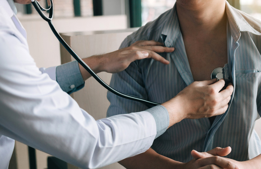 A person in a white jacket is using a stethoscope to listen to the heartbeat of person with a loosely buttoned striped shirt.