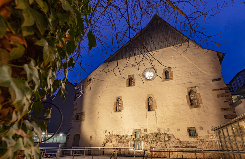 Photograph of a synagogue taken at night. A tree frames the left side. The facade of the building is lit from below. A rosette window is visible against the dark blue sky.