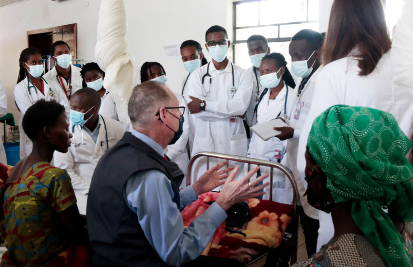 A line of women and men wearing white lab coats, medical masks, and stethoscopes stand facing a three people, including Paul Farmer, sitting on hospital beds. 