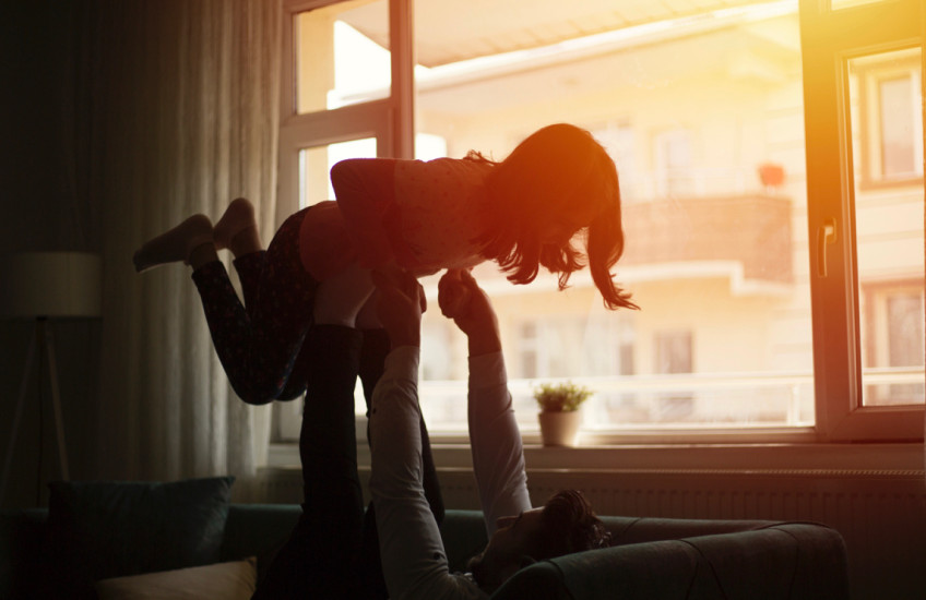 A father plays with his daughter backlit by sunset, seen through the window.