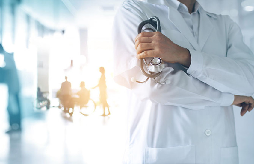 A physician with arms crossed stands in front of a blurry, overexposed background of a mostly empty waiting room.