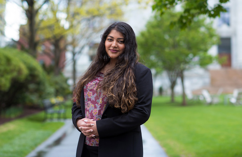 A woman with long, curly hair wearing a dark blazer and a flower-patterned shirt stands on a path between spring green bushes and lawn, an out of focus marble building in the background.