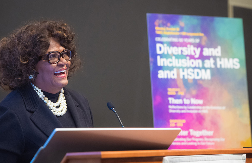 Alt Text:  A woman standing at a lectern, speaking in front of a colorful, out-of-focus poster