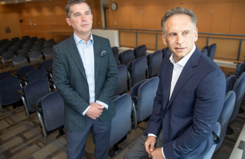 Two men in suits lean against chairs in an auditorium. 