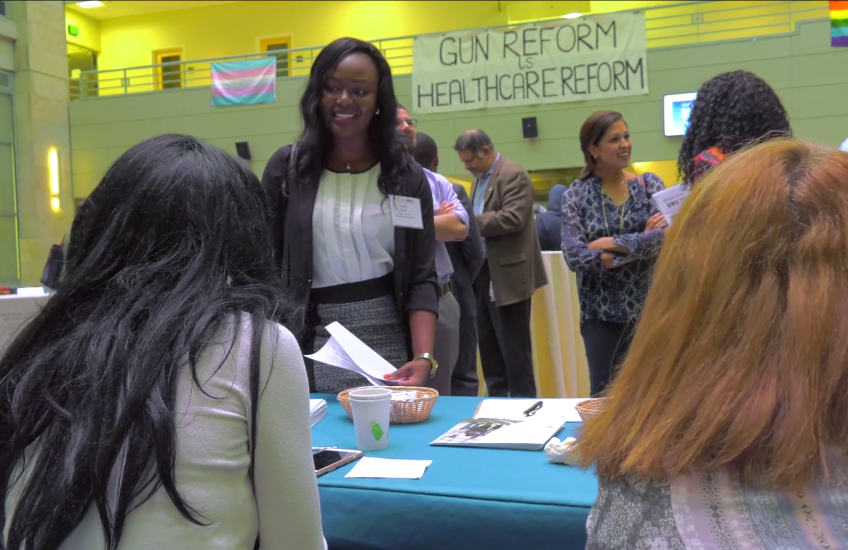 A student stands in front of a table, talking to two people seated at the table.