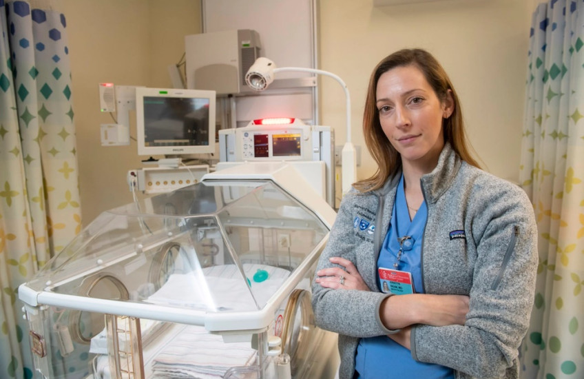 A woman in scrubs and a fleece stands with arms crossed next to a neonatal cradle.