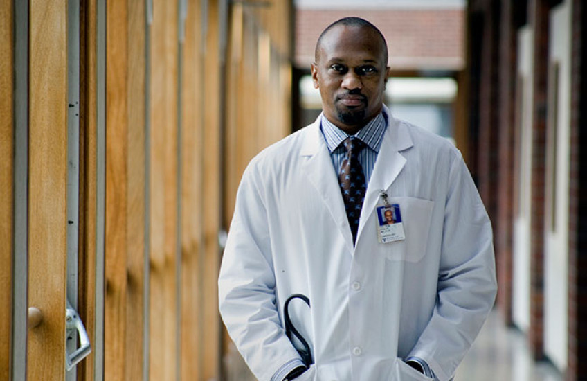 Stultz in a white coat with stethoscopes and hands in his pockets standing in a hallway