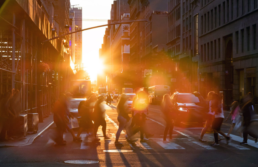 A group of people cross a street at a crosswalk, backlit by the glow of the sun and surrounded by a halo of rainbowed lens flare.