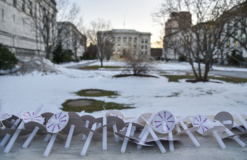 Unlit candles and paper holders line a stone wall at the edge of a snowy HMS Quadrangle, with Gordon Hall visible in background
